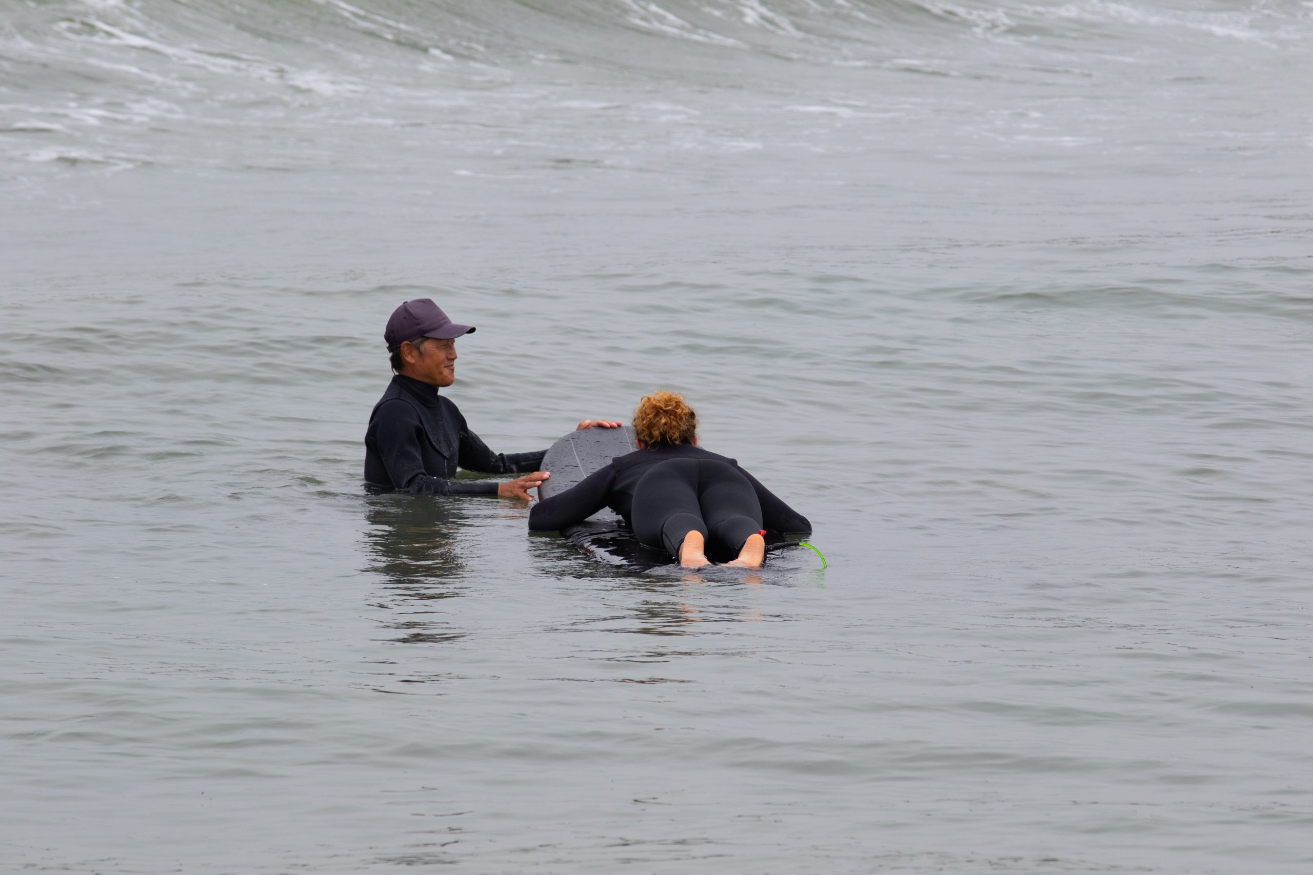 Young multiracial African American lady with amazing smile, freckles & frizzy hair & an Asian Japanese Surf Instructor having a surf lesson together in Chiba, Japan They are wearing black wetsuits.