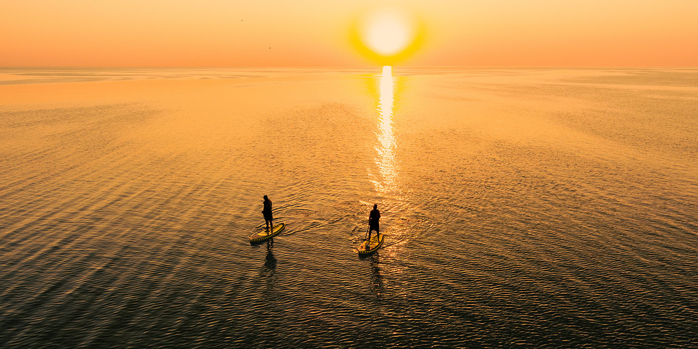 Aerial view of two people on stand up paddle boards on quiet sea at sunset. Warm summer beach vacation holiday.